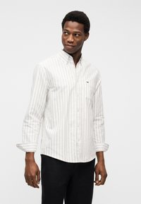 Άνδρας που φοράει λευκό πουκάμισο με ρίγες και κουμπιά, με τα μανίκια σηκωμένα, και μαύρο παντελόνι, κοιτάζει προς τα αριστερά σε απλό φόντο.