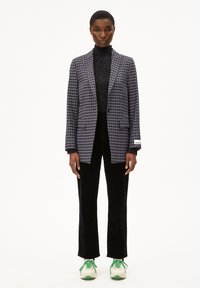 Geruite krijtjesblazer in marineblauw en wit, gecombineerd met een zwarte textuur colsjaal, zwarte fluwelen broek en groen-witte sneakers.