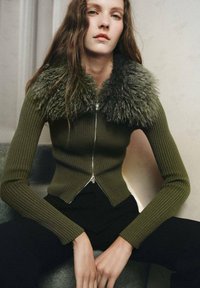 Olive green ribbed knit sweater with a faux fur collar, featuring a front zipper and fitted design, paired with black trousers.