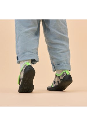 Kind trägt weiche Schuhe mit Tarnmuster, neongrüner Umrandung und weiße Socken, steht auf beigem Boden mit hochgekrempelten blauen Hosen.