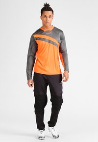 Langärmliges Sportshirt in Orange und Grau mit Rundhalsausschnitt, kombiniert mit schwarzen gepolsterten Hosen und weißen Sportschuhen.