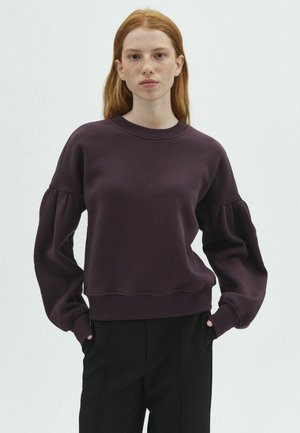 Giovane donna con capelli rossi che indossa un maglione viola scuro a maniche a sbuffo e pantaloni neri, in piedi con le mani in tasca davanti a uno sfondo semplice.