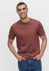 Kortærmet T-shirt med rund hals i rustfarve, lavet af bomuld. Modellen har jeans i denim på, står med hænderne i lommerne, neutral baggrund.