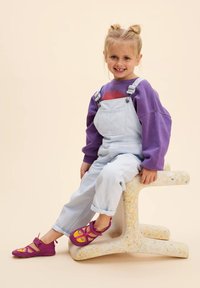 Purpurowa bluza, jasnoniebieskie ogrodniczki oraz żywo różowo-żółte buty. Dziecko siedzi na teksturowanym białym stołku z kolorowymi plamkami.