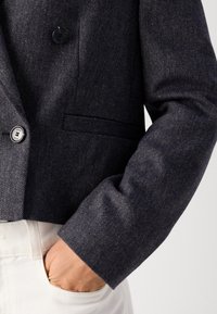 Blazer gris foncé texturé avec des boutons, porté sur un pantalon blanc, avec une main partiellement dans la poche.