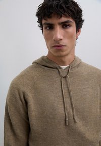 Jonge man met krullend donker haar in een beige gebreide hoodie met koordjes, tegen een effen lichtgrijze achtergrond.