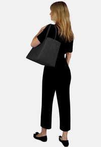 Schwarze geflochtene Tragetasche mit doppelten Griffen, rechteckiger Form, glatter Textur und festem Boden. Person in schwarzer Kleidung trägt sie über der Schulter.