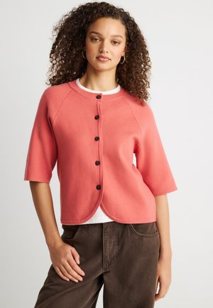 Mujer con cabello rizado que lleva un cárdigan coral abotonado sobre una camisa blanca y pantalones marrones, posando con una mano en el bolsillo frente a un fondo liso.