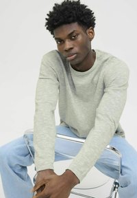Šviesiai pilkas, ilgomis rankovėmis marškinėliai su tekstūruotu trikotažu, derinami su šviesiai mėlynomis džinsinėmis kelnėmis. Modelis sėdi ant skaidraus kėdės, rankos uždėtos viena ant kitos.