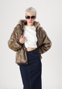 Ψεύτικη γουν jacket σε καφέ χρώμα με υφαντό σχέδιο, κουκούλα και φερμουάρ, συνδυασμένη με λευκή cropped μπλούζα και σκούρα τζιν φούστα.