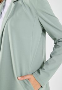 Light green jacket made of smooth fabric, featuring a tailored fit, long sleeves, and a side pocket. Seam details are visible.