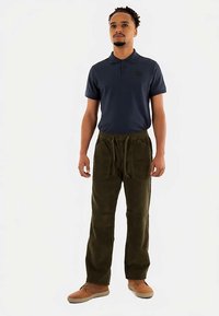 Mannen poloshirt in marineblauw met korte mouwen, gecombineerd met olijfgroene ribfluwelen broek met elastische tailleband en zijzakken. Bruine schoenen.