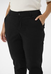 Cilvēks ar melniem biksēm un baltu krekls, vienu roku ielicis kabatā, bet otra roka mierīgi pie sāniem.