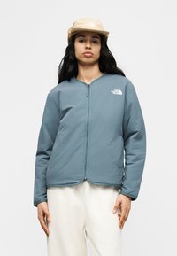 Άτομο που φορά μπλε ζακέτα με φερμουάρ και μπεζ καπέλο, στέκεται μπροστά σε απλό ανοιχτόχρωμο φόντο με τα χέρια χαλαρά δίπλα στο σώμα.