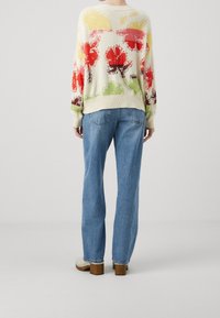 Pull en maille avec un motif floral en rouge, jaune et vert sur un fond crème, associé à un jean bleu à coupe décontractée et des sabots beiges.