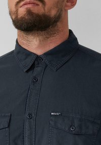Camicia blu navy con colletto a punta, due tasche frontali e un piccolo etichetta "Elevated Casual". Tessuto a finitura liscia.