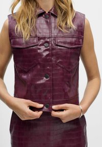 Chaleco de cuero sintético brillante de color púrpura, con un patrón de cocodrilo texturizado, diseño sin mangas y dos bolsillos en el pecho con botones negros.