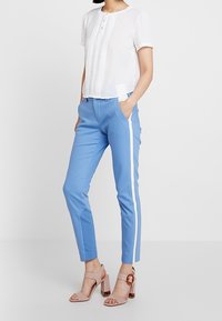 Witte blouse met korte mouwen en knoopsluiting, gecombineerd met blauwe taps toelopende broek met witte zijstrepen. Beige haksandalen maken de look compleet.