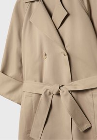 Trench-coat beige avec larges revers, boutons croisés, poches avant et ceinture en tissu nouée à la taille.
