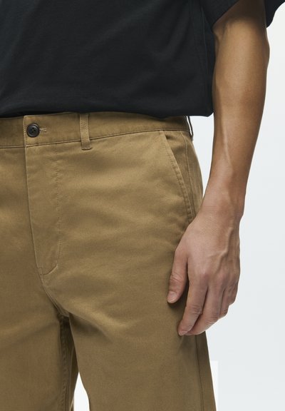 Pantalon en coton beige à coupe droite, avec une poche avant, une fermeture à bouton et des détails de couture subtils. Associé à une chemise noire.