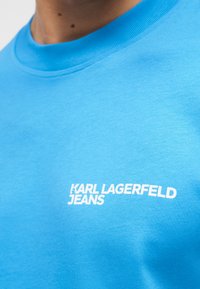 Άτομο που φοράει φωτεινό μπλε μπλουζάκι με στρογγυλή λαιμόκοψη και λευκό λογότυπο "KARL LAGERFELD JEANS" στο στήθος.