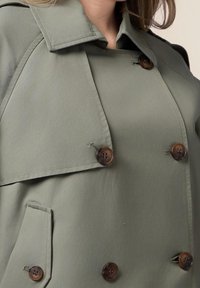 Olive green trench coat with a double-breasted design. Features large brown buttons, a notched collar, and side pockets. Smooth texture.
