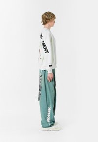 Sudadera de manga larga blanca con texto y gráficos negros en las mangas, combinada con jeans sueltos en teal claro con acentos laterales y etiquetas.