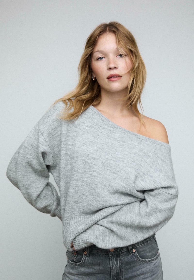Pull gris surdimensionné avec un design dénudé d'épaule, doté de poignets et d'ourlet côtelés. Associé à un jean bleu délavé taille haute.