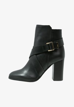 Czarny skórzany botek za kostkę na słupku z ozdobnym paskiem i złotym klamrą na białym tle.