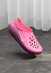 Svetloružová obuv na slip-on s ventilačnými otvormi a zúbkovanou podrážkou, vystavená vo vzduchu na šedej podlahe a v pozadí s bielou záclonou.