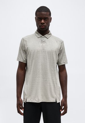 Bărbat purtând un tricou polo cu mânecă scurtă de culoare gri deschis, cu un logo mic pe piept și pantaloni negri, stând în fața unui fundal alb simplu.