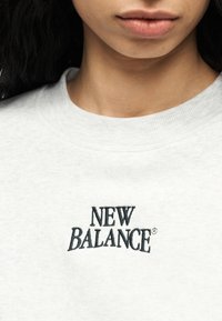 Κοντινό πλάνο ενός ατόμου που φορά ανοιχτό γκρι φούτερ με κεντημένο το «NEW BALANCE» με μαύρα γράμματα στο στήθος.