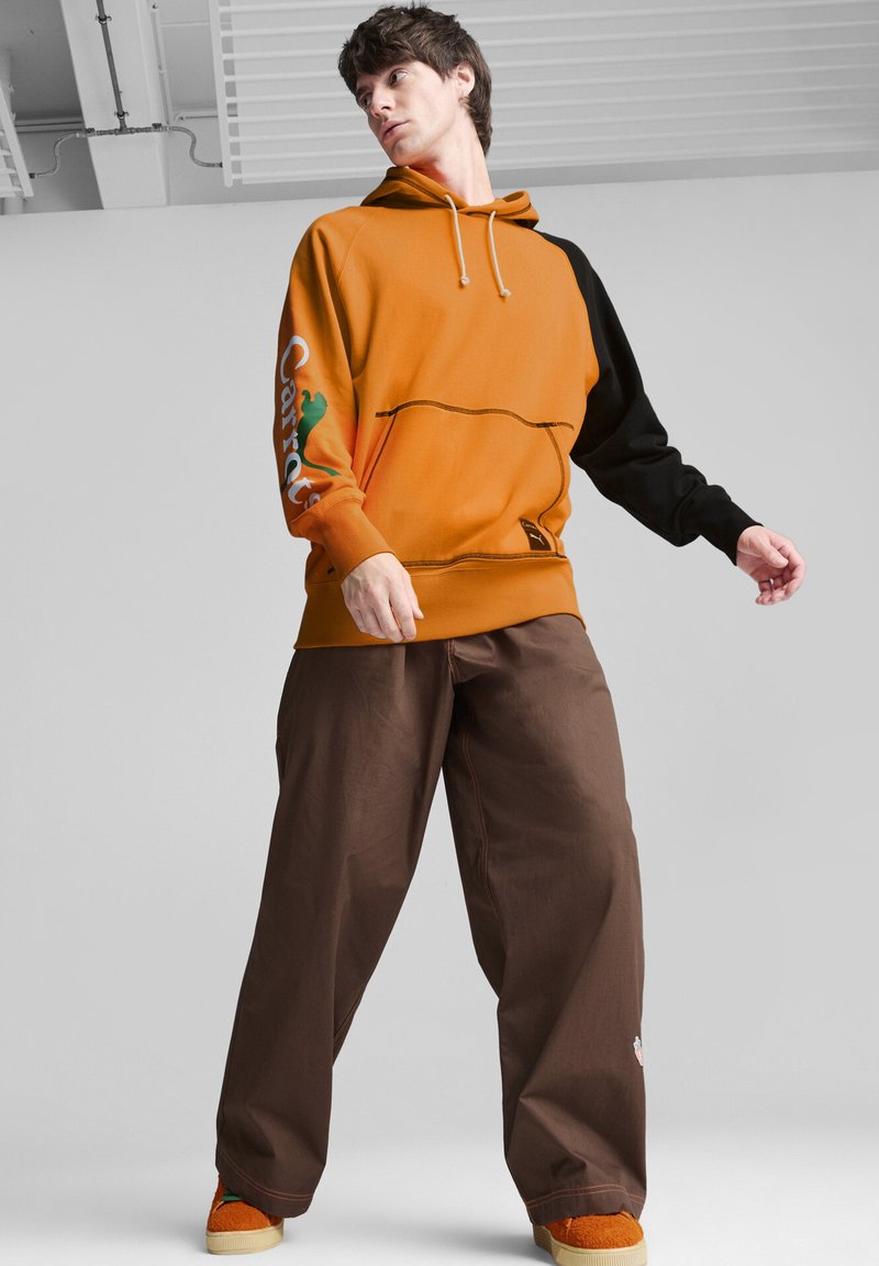 Felpa arancione e nera con cappuccio regolabile, tasca a marsupio e dettaglio del logo. Abbinata a pantaloni marroni larghi e scarpe arancioni.