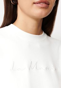 Biała bawełniana koszulka z okrągłym dekoltem i haftem "La Manis" w jasnoszarym kolorze. Zbliżenie na materiał i szwy.