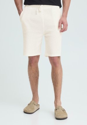 Weiße Strickshorts mit Kordelzug-Taschen, getragen von einer Person, die beige Slip-on-Schuhe vor einem weißen Hintergrund trägt.