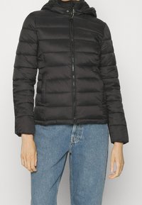 Czarna kurtka puchowa z zamkiem z przodu, kapturem i pikowaną fakturą. Posiada elastyczne mankiety oraz dopasowany krój. Nosi ją się na niebieskich dżinsach.