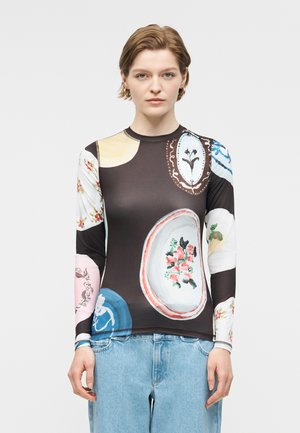 Γυναίκα που φοράει μαύρη μπλούζα με μακριά μανίκια, διακοσμημένη με πολύχρωμα αφηρημένα σχέδια λουλουδιών και πλακέ μοτίβα, συνδυασμένη με ανοιχτό μπλε τζιν.