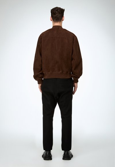 Ein Mann steht mit dem Rücken zur Kamera und trägt eine braune Wildlederjacke, schwarze Hosen und schwarze Schuhe vor einem schlichten hellen Hintergrund.