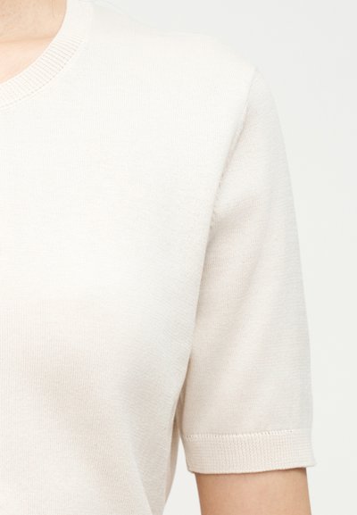 Κοντινό πλάνο ενός ατόμου που φοράει κοντομάνικο, ανοιχτόμπεζ πλεκτό τοπ με στρογγυλή λαιμόκοψη και ριμπ μανσέτες.