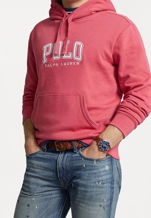 Roter Hoodie mit weißem "POLO RALPH LAUREN"-Schriftzug, Kängurutasche, getragen mit zerrissenen blauen Jeans, geflochtenem braunem Gürtel und einer blauen Uhr.