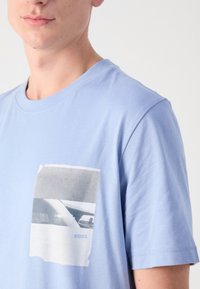 Jeune homme portant un T-shirt bleu clair avec une impression photo en noir et blanc et le mot "BOSS" sur la zone de la poche poitrine.