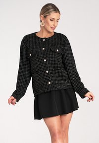 Czarna pikowana kurtka w kratę, z srebrnymi guzikami i dwoma kieszeniami na piersi. Noszona na czarnej spódnicy, z długimi rękawami.