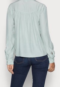Blouse verte clair à manches longues avec un dos froncé et des poignets à boutons, associée à un jean bleu foncé. Tissu doux et fluide.
