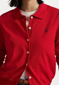 Cardigan rouge en tricot avec col, doté d'une fermeture à boutons et d'un petit logo bleu marine. Porté par-dessus une chemise blanche, superposé pour un effet de texture.
