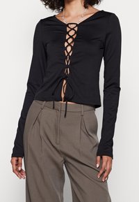 Top nero a maniche lunghe con scollo a V profondo, caratterizzato da una chiusura lacci e dettagli arricciati regolabili. Abbinato a pantaloni sartoriali a vita alta.