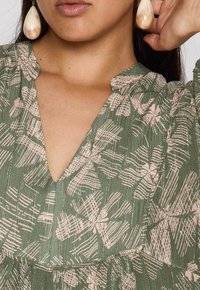 Vrouw in groene jurk met patroon en V-hals, met grote crèmekleurige druppeloorbellen, hand rustend bij het oor.