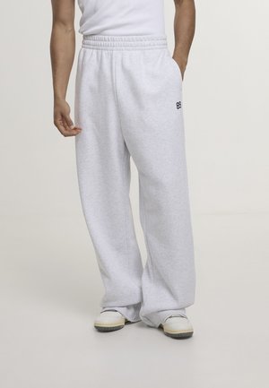 Personne portant un pantalon de survêtement ample gris clair avec une taille élastique et des baskets blanches, debout devant un fond uni clair.