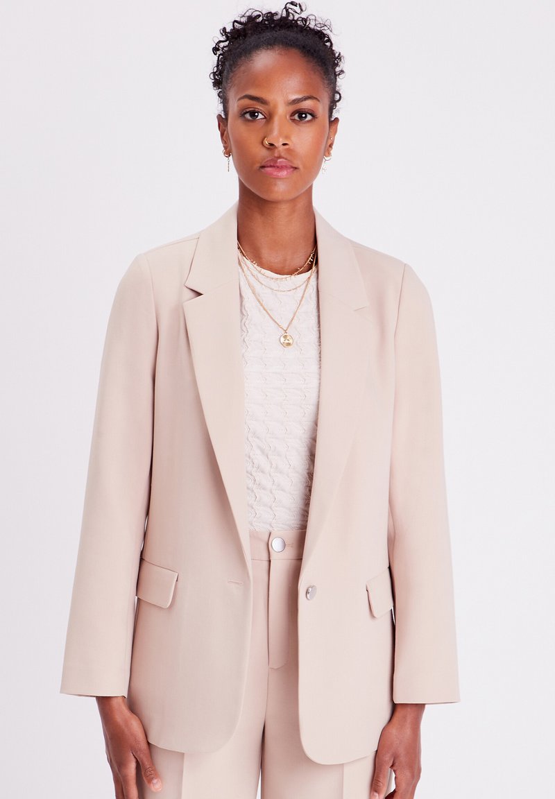 Light beige tailored blazer with a single button, notched lapels, and two front pockets. Worn over a textured cream top.