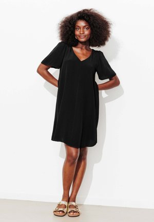 Robe noire à manches courtes avec un col en V, coupe ample et longueur aux genoux. Le modèle porte des sandales dorées claires avec un design à deux boucles.