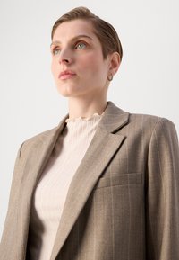 Braunes Blazer mit Nadelstreifen, breiten Revers und einer aufgesetzten Tasche, über einem strukturierten hellen Cremetop mit rüschigem Ausschnitt getragen.